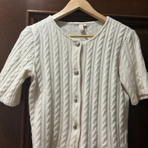 Vit stickad blus med silver knappar från Nelly storlek S - Säljer en vit stickad blus från Nelly med korta ärmar och snygga silverfärgade knappar framtill. Blusen har ett klassiskt kabelstickat mönster och rund halsringning. 