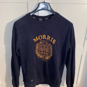 Svart sweatshirt från Morris Rowing Club - Svart sweatshirt från Morris Rowing Club med guldfärgat tryck och emblem framtill. Tröjan har rund halsringning, långa ärmar och är tillverkad i mjuk bomull. Perfekt för en preppy och sportig look.