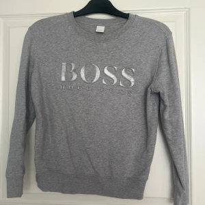 Grå sweatshirt från Hugo Boss - Snygg grå sweatshirt från Hugo Boss med silverfärgat BOSS-tryck på bröstet. Tröjan har rund halsringning och långa ärmar. Perfekt för en stilren och avslappnad look. Köpt från zalando. Köpt för längesen men knappt använd