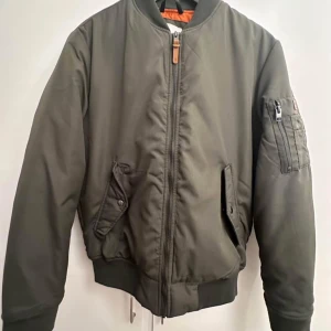 Olivgrön bomberjacka Jack & Jones - Snygg olivgrön bomberjacka från Jack & Jones med orange quiltat foder. Jackan har ribbade muddar, dragkedja framtill, fickor med tryckknappar och en extra ficka med dragkedja på ärmen. Jackan är knappt använd.