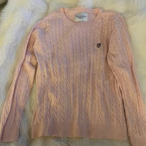 Ljusrosa kabelstickad tröja Bondelid - Supersöt ljusrosa kabelstickad tröja från Bondelid med rund hals och diskret logga på bröstet. Tröjan har långa ärmar och ribbade muddar vid ärmslut och nederkant. Perfekt för lager-på-lager och chill dagar.