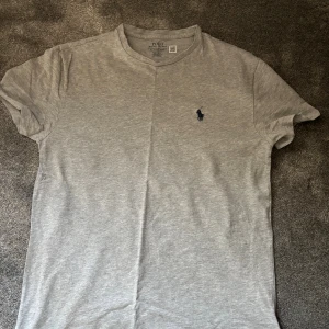 Grå t-shirt från Polo Ralph Lauren XS - Snygg grå t-shirt från Polo Ralph Lauren i klassisk modell. T-shirten har korta ärmar, rund halsringning och den ikoniska mörkblå loggan broderad på bröstet. Tillverkad i mjuk bomull för en skön känsla och enkel stil. Skicket är 9,5/10 och använd fåtal gånger! 
