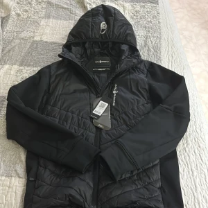 Svart cardigan från Sail Racing - Svart pufferjacka från Sail Racing med huva och dragkedja framtill. Jackan har quiltade partier på bröst och mage, samt släta ärmar. Logga på bröstet och huvan. Tillverkad i syntetmaterial, perfekt för kyliga dagar.