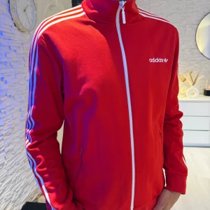 Röd Adidas track jacket med vita ränder - Säljer en röd Adidas track jacket i storlek M med klassiska vita ränder längs ärmarna och vit dragkedja. Jackan har hög krage, två fickor med dragkedja och Adidas-logga på bröstet. Perfekt för en sportig och stilren look. Passar bra som oversize modell.