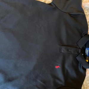 Svart pikétröja från Polo Ralph Lauren - Klassisk svart pikétröja från Polo Ralph Lauren i bomull med röd broderad logga på bröstet. Tröjan har krage, knappar framtill och korta ärmar. Perfekt för en clean och stilren look.