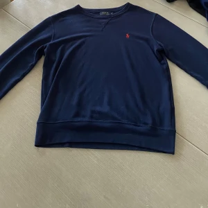 Mörkblå tröja från Polo Ralph Lauren - Klassisk mörkblå tröja från Polo Ralph Lauren med rund halsringning och broderad röd logga på bröstet. Tröjan har långa ärmar och är tillverkad i mjuk bomull, perfekt för en stilren och avslappnad look.