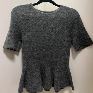 Grå stickad topp med volang - Säljer en grå stickad topp med korta ärmar och rund hals. Toppen har en snygg volangkant nedtill som ger en cool och feminin touch. 