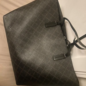 Stor svart shoppingväska med rutmönster - Snygg och rymlig shoppingväska i svart med diskret rutmönster. Väskan har långa handtag och är tillverkad i ett tåligt material som liknar skinn. Perfekt för dig som vill ha plats för allt du behöver under dagen.