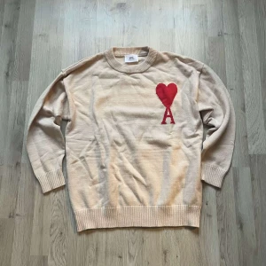 Beige stickad tröja från Ami Paris - Säljer en beige stickad tröja från Ami Paris med röd hjärtlogga och bokstaven A på bröstet. Tröjan har rund hals, ribbade muddar och är långärmad. Perfekt för dig som gillar stilrena plagg med en twist.