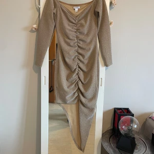 Glittrig asymmetrisk klänning NLY One - Glittrig beige långklänning från NLY One med asymmetrisk nederkant och rynkade detaljer framtill. Klänningen har lång ärm, v-ringning och figurnära passform. Perfekt för dig som vill sticka ut med en unik och trendig look.