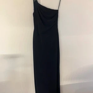 Marinblå långklänning oneshoulder - Säljer en marinblå långklänning med oneshoulder och smal axelrem. Klänningen har snygga draperingar och dekorativa detaljer vid axeln. Den är figurnära och elegant, perfekt för dig som vill sticka ut med en stilren look.