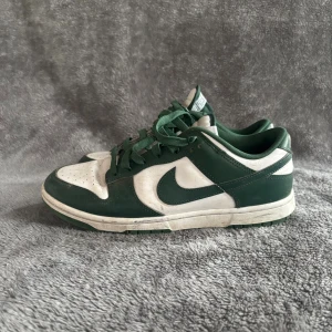 Nike Dunk Low grön/vit sneakers - Snygga Nike Dunk Low sneakers i grönt och vitt med klassisk swoosh-logga på sidan. Skorna har snörning, platt sula och är tillverkade i skinn och syntet. Perfekta för dig som gillar streetwear och vill sticka ut med en ikonisk modell. Storlek 42