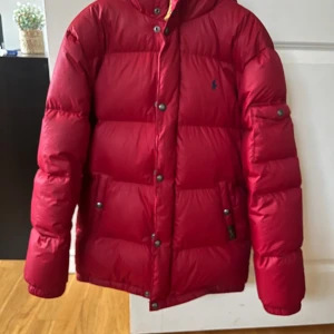 Röd dunjacka från Polo Ralph Lauren - Riktigt fet röd dunjacka från Polo Ralph Lauren med huva och diskret logga på bröstet. Jackan är vadderad, har knappar och dragkedja framtill samt fickor med tryckknappar. Perfekt för kalla dagar när du vill hålla stilen. Jackan är i storlek kids L vilket motsvarar xs-s