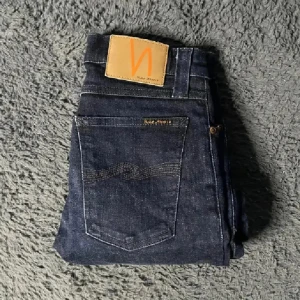 Mörkblå jeans från Nudie Jeans - Snygga mörkblå jeans från Nudie Jeans med klassisk femficksdesign och hög midja. Jeansen har Nudie-loggan på bakfickan samt en läderpatch i midjan. Perfekt passform och stilren look.