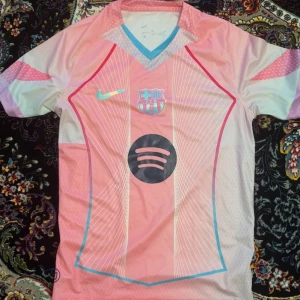 Rosa FC Barcelona fotbollströja Nike - Säljer en snygg FC Barcelona fotbollströja från Nike i rosa med ljusblå och vita detaljer. Tröjan har korta ärmar, V-ringad krage med blå kant och tryck med Spotify-logga på bröstet. Materialet är lätt och andas, perfekt för match eller träning.