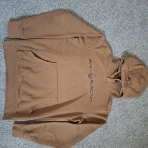 Beige hoodie från Sail Racing - Nypris 1500 kr.  Säljer en snygg beige hoodie från Sail Racing med tryckt logga på bröstet. Tröjan har huva med snörning och en stor magficka. Perfekt för dig som gillar en clean och stilren look.