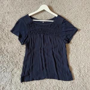 Mörkblå T-shirt med broderad detalj - Mörkblå blus från Laura Ashley med korta ärmar och rund halsringning. Ovanför bysten finns en snygg broderad detalj som ger toppen en unik look. Tillverkad i mjukt tyg som faller fint. Nästan aldrig använd💓