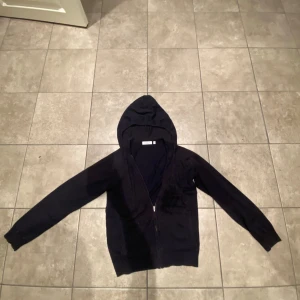 Mörkblå hoodie från RYVLS Stockholm - Mörkblå hoodie från RYVLS Stockholm med dragkedja framtill och snörning i huvan. Tröjan har långa ärmar, ribbade muddar och två fickor på framsidan. Perfekt för dig som gillar enkel och stilren design.