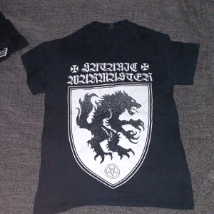 Svart Satanic Warmaster t-shirt - Svart t-shirt med stort tryck fram och bak från Satanic Warmaster. Framsidan har en sköld med en varg och pentagram, samt bandets logga i vit gotisk stil. Baksidan har en stor vit symbol. Klassisk passform och rund hals.