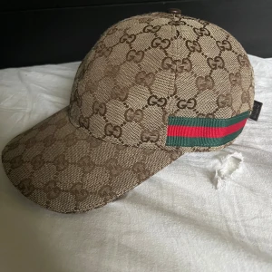 Gucci beige keps med GG-mönster - Snygg beige keps från Gucci med klassiskt GG-mönster över hela kepsen. På sidan finns den ikoniska gröna och röda randen. Justerbar rem baktill i brunt skinn. Perfekt för dig som vill ha en lyxig och trendig accessoar.