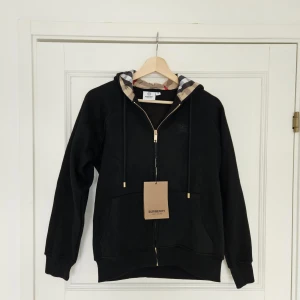 Svart Burberry hoodie med dragkedja - Svart hoodie från Burberry med klassiskt rutigt foder i huvan och guldfärgad dragkedja. Tröjan har broderad Burberry-logga på bröstet, snörning i huvan och två fickor framtill. Materialet är mjuk bomull. Passformen är ganska liten så jag skulle rekommendera att köpa den om du har storlek S.