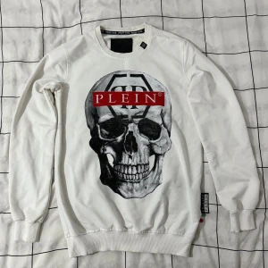 Vit sweatshirt med döskalle från Philipp Plein - Säljer en vit långärmad sweatshirt från Philipp Plein med stor döskalleprint och röda PLEIN-loggor både fram och bak. Tröjan har rund hals, ribbade muddar och coola grafiska detaljer. Materialet är mjuk bomull som känns skönt mot huden.