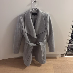 Grå kappa med bälte från Vila - Snygg grå kappa från Vila i storlek S med bred krage och knytbälte i midjan. Jackan har en stilren design med långa ärmar och dubbla knappar framtill. Perfekt för dig som gillar klassisk look med modern touch. Kappan är knä lång.