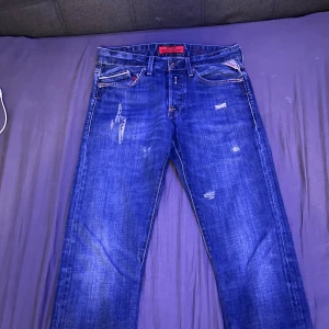 Blå Replay jeans med slitningar, W30 L32 - Säljer ett par snygga blå jeans från Replay med coola slitningar och detaljer. Klassisk femficksmodell med röd Replay-logga på fickan och etikett i midjan. Jeansen har raka ben och är tillverkade i slitstarkt denimtyg. Perfekt för dig som gillar streetstyle.