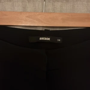 Svarta bootcut-byxor från Bik Bok - Svarta kostymbyxor från Bik Bok i storlek 34. Modell PW Vera. Byxorna har en klassisk siluett med utsvängda ben och midja med knappstängning. Materialet känns mjukt och följsamt. Följer med reservknapp 