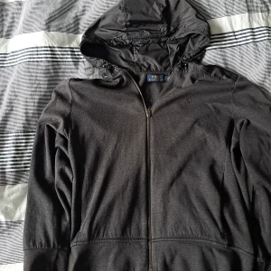 Svart hoodiejacka från Polo Ralph Lauren - Svart hoodiejacka från Polo Ralph Lauren med dragkedja och broderad logga på bröstet. Jackan har huva och detaljer i syntetmaterial på huva och nederkant. Perfekt för dig som gillar stilren och sportig look.