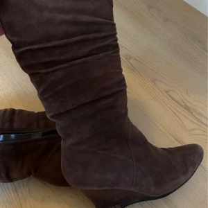 Bruna mockastövlar med kilklack - Snygga bruna stövlar i mocka med kilklack och spetsig tå. Stövlarna har dragkedja på sidan och är fodrade för extra värme. Perfekta för höst och vinter när du vill ha både stil och komfort.