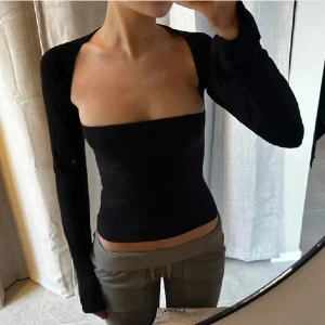 Svart croppad bolerotopp - Säljer en svart croppad bolerotopp med lång ärm och öppen axel. Toppen har en tight passform och är axelbandslös framtill, vilket ger en modern och edgy look. Perfekt för dig som gillar minimalistisk stil och vill sticka ut.