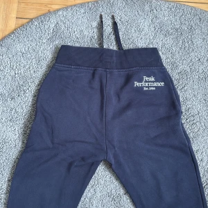 Mörkblå mjukisbyxor Peak Performance - Snygga mörkblå mjukisbyxor från Peak Performance med broderad logga på höften. Byxorna har ribbad midja med snörning, muddar vid benslut och två sidofickor. Perfekta för chill eller träning, riktigt sköna och stilrena.