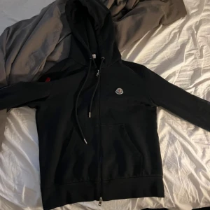Moncler hoodie  - Svart hoodie från Moncler med huva, snörning och hel dragkedja. Tröjan har Moncler-logga på bröstet och en snygg detalj med röd och blå rand längs dragkedjan. Tillverkad i mjuk bomull, perfekt för chill dagar.