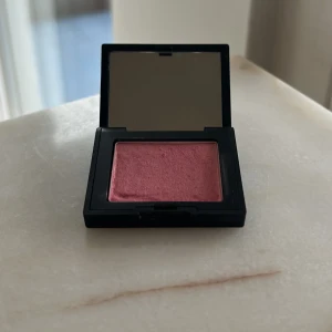 NARS rouge - Ikoniskt rouge från NARS i nyansen Orgasm i travel size. En persikorosa färg med guldskimmer. Kommer i en svart, matt dosa med spegel i locket. Perfekt för att ge kinderna en fräsch glow och naturlig rodnad.