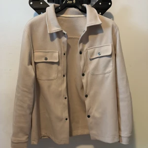 Beige overshirt med fickor - Snygg beige overshirt med klassisk krage och tryckknappar framtill. Jackan har två stora bröstfickor med lock och knapp, samt långa ärmar. Materialet är mjukt och har en fleece-liknande känsla, perfekt för lager-på-lager. Enkel och clean stil som funkar till allt.