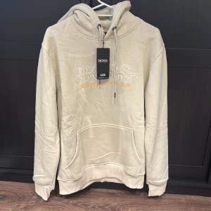 Beige hoodie från Hugo Boss - Snygg beige hoodie från Hugo Boss med broderad logga i vitt och orange framtill. Tröjan har huva med snörning, stor magficka och ribbade muddar. Perfekt för en chill och stilren look.
