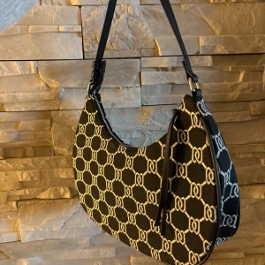 Svart och vit axelväska med mönster - Snygg svart axelväska med vit kedjeliknande mönster över hela väskan. Väskan har en böjd form och ett kort axelband i svart. Stängs med dragkedja och är tillverkad i ett tygmaterial med struktur. Perfekt för dig som gillar trendiga accessoarer.