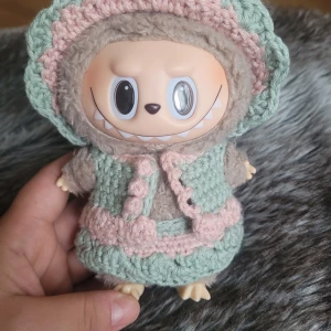 Handvirkade labubu kläder. - Supersöta handvirkade kläder till Labubu i mintgrönt och ljusrosa. Setet består av klänning, väst och hatt med vågig kant och dekorativa detaljer. Perfekt för att ge dockan en gullig och unik look. Gjord i mjukt garn för extra mysfaktor.