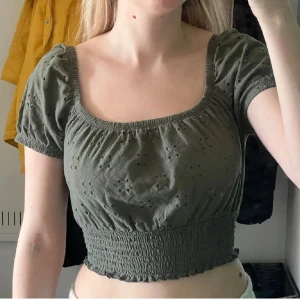 Olivgrön croppad topp från Hollister - Supersöt olivgrön croppad topp från Hollister med broderade detaljer och smockad midja. Toppen har korta puffärmar, fyrkantig urringning och små hålmönster i tyget. Perfekt för dig som gillar en feminin och trendig stil. Man kan använda den som ofshoulder också