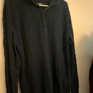 Svart kabelstickad pikétröja GANT - Svart kabelstickad pikétröja från GANT med klassisk krage och tre knappar framtill. Tröjan har lång ärm och diskret broderad logga på bröstet. Materialet är mjukt och stickat, perfekt för dig som gillar stilren och tidlös design.