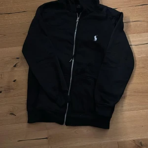 Svart hoodie från Polo Ralph Lauren - Svart hoodie från Polo Ralph Lauren med vit broderad logga på bröstet. Tröjan har huva, hel dragkedja och två fickor framtill. Tillverkad i mjuk bomull, perfekt för chill dagar. Klassisk och stilren design med ribbade muddar vid ärmslut och nederkant. Helt ny och jag säljer den för billigt för att jag vill ha pengar till en del som fattas på min EPA