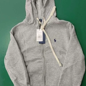 Grå hoodie från Polo Ralph Lauren - Grå hoodie från Polo Ralph Lauren med dragkedja och huva. Klassisk design med mörkblå logga broderad på bröstet och vita snören i huvan. Tillverkad i mjuk bomullsmix, perfekt för chill dagar. Långärmad och med fickor framtill.