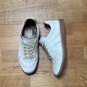 Maison Margiela vita sneakers i skinn - Maison Margiela sneakers i vitt skinn med klassisk rund tå och platt gummisula i brunt. Snygga paneldetaljer och snörning framtill. Ikonisk Margiela-etikett på plösen. Perfekt för dig som gillar minimalistisk och clean stil.