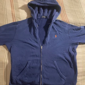 Blå hoodie från Polo Ralph Lauren - Säljer en blå hoodie från Polo Ralph Lauren med dragkedja och huva i storlek L. Jag säljer då jag vill rensa lite i garderoben och behöver lite extra pengar.