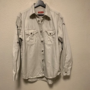 Beige skjorta från Replay i bomull - Stilren beige skjorta från Replay med klassisk krage, två bröstfickor med lock och knappar. Skjortan har långa ärmar med knappdetaljer och är tillverkad i mjuk bomull. Perfekt för en avslappnad men snygg look.