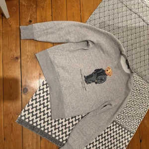 Grå Polo Bear sweatshirt från Ralph Lauren - Grå sweatshirt från Ralph Lauren med ikoniska Polo Bear-trycket framtill. Tröjan har rund hals, ribbade muddar och mjuk insida. Perfekt för dig som gillar streetstyle och vill ha något bekvämt men ändå snyggt.