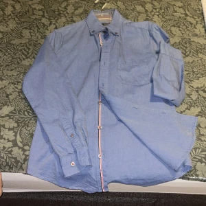 Blå slim fit skjorta från Dressmann - Snygg blå skjorta från Dressmann i slim fit-modell. Skjortan har långa ärmar, klassisk krage och coola detaljer med rödvit rand längs knappraden. Perfekt för dig som gillar stilrena plagg med en twist.