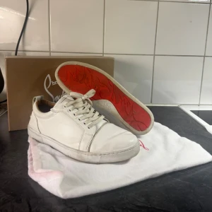 Vita sneakers från Christian Louboutin - Säljer ett par stilrena vita sneakers från Christian Louboutin med klassisk röd sula och snörning. Skorna är i skinn och har en rund tå samt platt sula. Perfekta för dig som vill ha något exklusivt och snyggt till din outfit.
