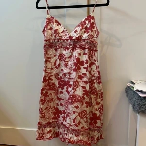 Rödvit blommig sommarklänning bomull - Supersöt röd och vit blommig kortklänning i 100% bomull med smala axelband och v-ringning. Klänningen har smockad midja och volangkant nertill, perfekt för varma dagar. Lätt och luftig känsla, lining i bomull. Snygg till sneakers eller sandaler!